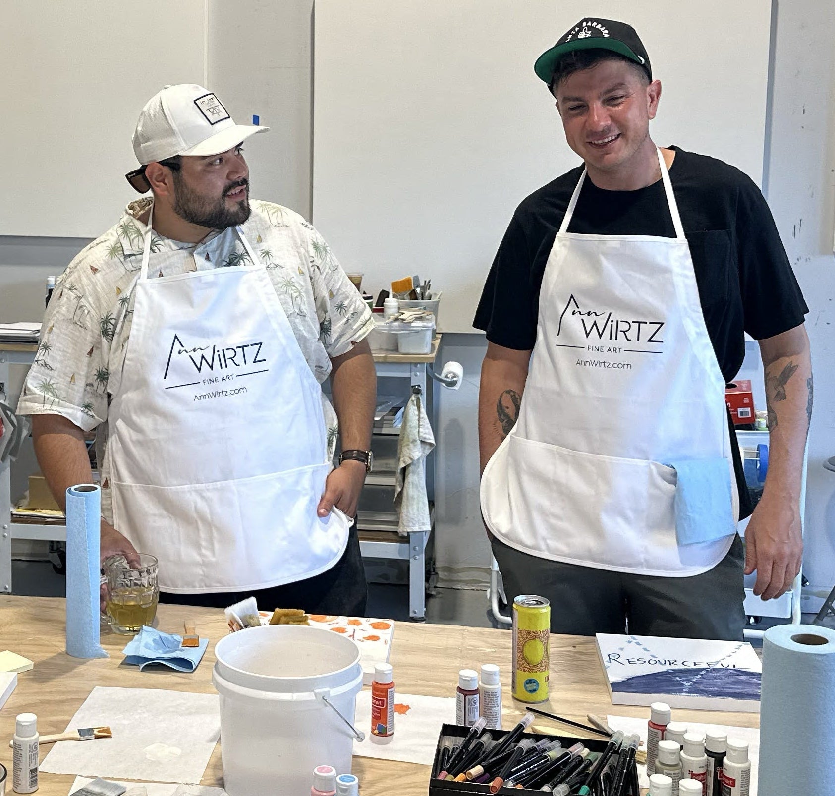 Three people wearing Wirtz aprons standing around a table with art supplies in a studio setting.