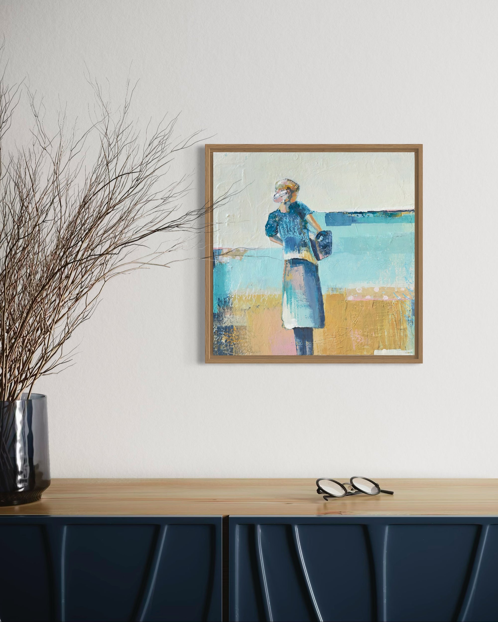 Framed artwork on a wall above a blue cabinet with a vase of branches.